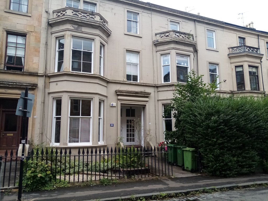 a two storey tenement with off-white render, spiked
     railings, bay windows either side of the recessed doorway